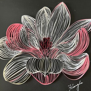 May include: A paper quilling art piece of a flower with white, pink, and dark red petals on a black background. The flower is made of layered paper strips, creating a 3D effect. The artwork is signed in the bottom right corner.