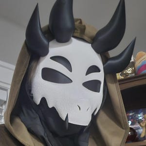 May include: A brown hooded cloak worn over a black garment, featuring a white mask with black eye cutouts and four black horns. A silver-toned brooch shaped like a dragon with green accents is pinned to the front of the cloak near a metal ring closure.