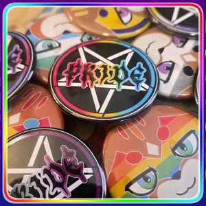 May include: A collection of circular pin-back buttons featuring various designs, including a black pentagram with the word PRIDE in rainbow lettering, and stylized cartoon animal faces with rainbow-colored accents and bold graphic outlines.