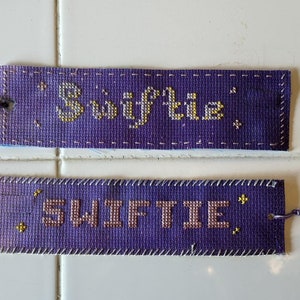 May include: Two purple fabric bookmarks with the word "Swiftie" embroidered in gold thread. The bookmarks have a tassel on one end.
