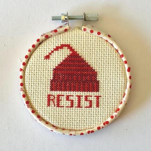 以下が含まれることがあります： 赤い建物のデザインと「RESIST」の文字が赤色で刺繍された、小さくて丸いクロスステッチの刺繍枠。 赤い水玉模様の生地で縁取られています。 刺繍は金属製のネジで固定されています。