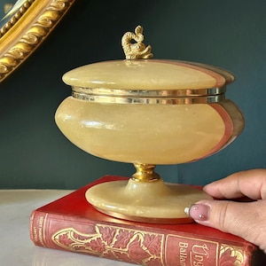 Pode incluir: Uma taça com tampa feita de pedra translúcida amarelo-pálido, com uma borda de metal dourado e um pequeno remate em forma de golfinho dourado na tampa. A taça é suportada por uma base de pedestal feita da mesma pedra amarelo-pálido.