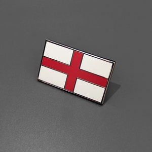 Könnte beinhalten: Eine silberne Anstecknadel mit einem roten Kreuz auf weißem Hintergrund. Die Anstecknadel ist eine Darstellung der englischen Flagge.