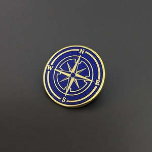 Puede incluir: Un pin de brújula de oro con fondo de esmalte azul. La brújula apunta al norte, sur, este y oeste.