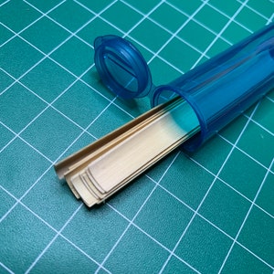 May include: A blue plastic container with a lid, containing a stack of thin, gold-colored metal strips.