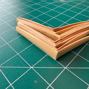 May include: A stack of thin, light brown, rectangular pieces of wood folded in a V-shape on a green cutting mat.
