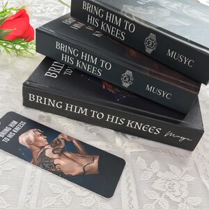May include: Three black books stacked, titled "BRING HIM TO HIS KNEES" in white lettering. A bookmark with the same title and a male illustration is in front. The books have a watch design and the word "MUSYC".
