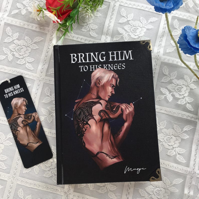 May include: A book and bookmark with the title "BRING HIM TO HIS KNEES" on a black background. The book and bookmark feature an illustration of a shirtless man with tattoos. The book has gold corner protectors.