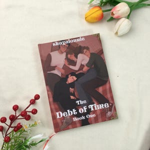 May include: A book titled "The Debt of Time, Book One" with an illustration of four people. The cover art features a maroon background with the author's name, "skayalonnie," at the top. A red ribbon bookmark is visible.