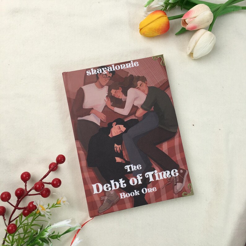 May include: A book titled "The Debt of Time, Book One" with an illustration of four people. The cover art features a maroon background with the author's name, "skayalonnie," at the top. A red ribbon bookmark is visible.