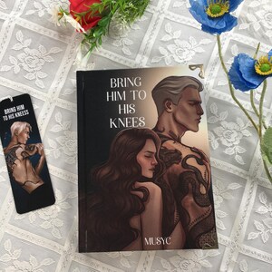 May include: A book and bookmark titled "Bring Him to His Knees." The book cover shows a couple, with the man's back covered in tattoos. The bookmark has the same design. The background is a white lace tablecloth.