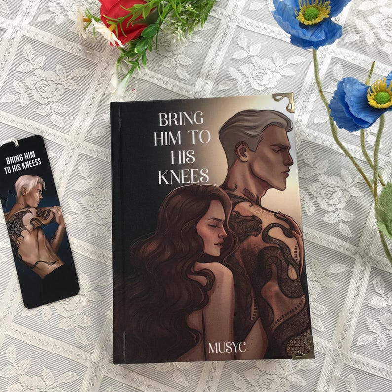 May include: A book and bookmark titled "Bring Him to His Knees." The book cover shows a couple, with the man's back covered in tattoos. The bookmark has the same design. The background is a white lace tablecloth.