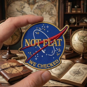 May include: A blue, circular patch with the NASA logo, featuring the text "NOT FLAT WE CHECKED" in gold lettering. The patch has a red swoosh and white stars, held in a hand against a backdrop of maps and globes.