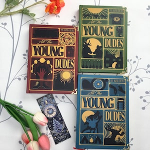 May include: A collection of hardcover books with the title "All The Young Dudes" in yellow lettering. The books are in burgundy, green, and blue, with decorative illustrations and gold corner accents. A matching bookmark is also present.