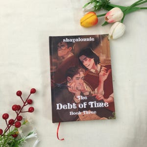 May include: A hardcover book titled "The Debt of Time Book Three" with an illustration of three people. The cover features the author's name, "shayalorrie". A red ribbon bookmark is visible.