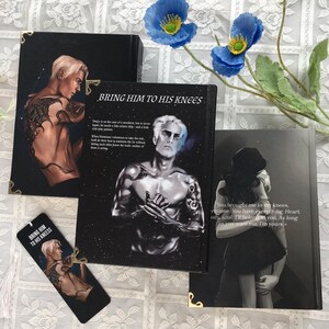 May include: A selection of books and a bookmark, all featuring illustrations and text. The book covers display a male figure with tattoos and the title "BRING HIM TO HIS KNEES." The bookmark also shows the title and a similar illustration. Blue flowers are visible.