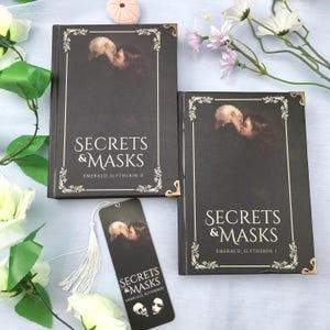 May include: Two hardback books and a bookmark with the title "Secrets & Masks" in white font. The books are black with gold corner accents and a decorative border. The bookmark has a tassel.