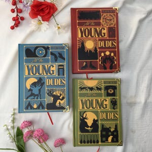 May include: Three hardcover books titled All The Young Dudes, each featuring distinct cover art in blue, red, and green. The covers display intricate graphic illustrations, gold-toned corner protectors, and ribbon bookmarks, arranged on a white fabric.