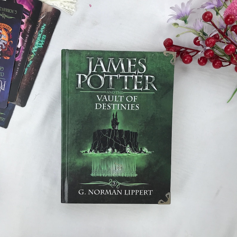 May include: A hardcover book titled "JAMES POTTER and the VAULT OF DESTINIES" by G. Norman Lippert. The cover is predominantly green with a fantasy illustration of a castle and water. Other books and flowers are in the background.