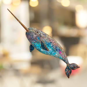 May include: A mosaic-style narwhal with a long, thin, pointed horn. The narwhal is made of blue, purple, and pink glass tiles with gold accents.