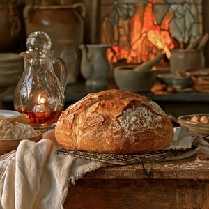 May include: A round, golden-brown loaf of crusty sourdough bread sits on a metal wire cooling rack. The bread has a scored, flour-dusted top and a rustic texture. A clear glass carafe with a faceted stopper and amber liquid is positioned nearby.
