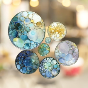May include: A set of six round, colorful, abstract art pieces with a bubble-like design. The pieces are made of resin and have a glossy finish. The colors are blue, green, yellow, and white.