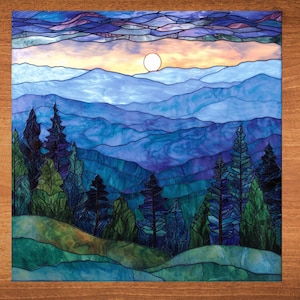 May include: Stained glass panel depicting a mountain landscape at sunset. The artwork features layers of blue mountains, green trees, and a yellow sun. The sky transitions from orange to purple and blue.