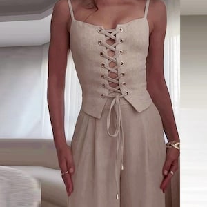May include: A beige linen two-piece outfit. The top is a corset-style camisole with spaghetti straps and a lace-up front. The matching high-waisted trousers have a wide leg. The outfit is made of a light, neutral-toned fabric.