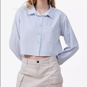 May include: A light blue, cropped button-down shirt with vertical stripes and a single chest pocket. The shirt has long sleeves and a classic collar. Paired with a beige cargo mini skirt.