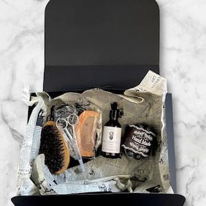 Puede incluir: Una caja de regalo negra con productos de aseo. En su interior, un cepillo de madera, un peine, unas tijeras, una botella de aceite y una pastilla de jabón, dispuestos sobre papel de periódico. La caja está abierta, mostrando el contenido.