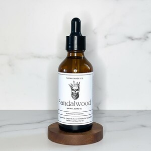 May include: A brown glass bottle of sandalwood natural beard oil with a black dropper top. The label features a skull with a crown and the text "Sandalwood". The bottle sits on a small, round wooden stand.