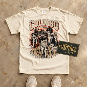 May include: A cream-coloured short-sleeve t-shirt featuring a graphic print of the television character Columbo. The design includes the text COLUMBO, a car, and three figures in a circular frame. A black rectangular sign reads VINTAGE T-SHIRT.