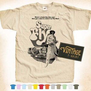 May include: A vintage t-shirt featuring a black and white image of a man in a long coat standing next to a car. The text "Super Fly" is in a large, stylized font above the image. The text "Never a dude like this one! He's got a plan to stick it to The Man!" is printed above the title. The text "Vintage T-Shirt" is printed in a black rectangle in the bottom right corner.