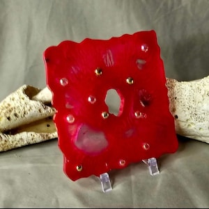 May include: A square, red, resin art piece with a hole in the center and gold accents. The piece is on a clear stand.