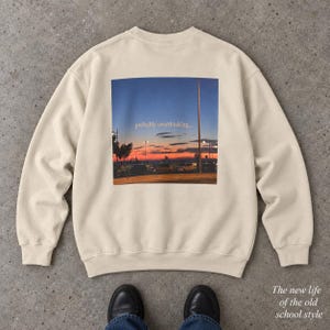 May include: A beige crewneck sweatshirt featuring a large square graphic on the back. The graphic depicts a sunset over a parking lot with the text "probably overthinking..." centred in the middle of the image. The sweatshirt has long sleeves.