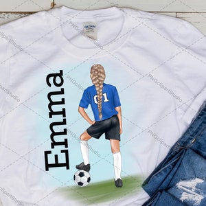 May include: White t-shirt with the name "Emma" printed in black on the left side. On the right side, a girl with blonde hair is wearing a blue soccer jersey with the number "01" on the back. She is holding a soccer ball and is standing on a green field.