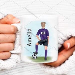 May include: White ceramic mug with a blue background and a cartoon illustration of a girl wearing a purple soccer uniform and holding a soccer ball. The text "Amelia" is written in black on the left side of the mug and the number "01" is on the girl's jersey.