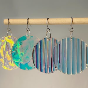 Puede incluir: Cuatro pares de pendientes redondos e iridiscentes colgados de una clavija de madera. Los pendientes tienen una variedad de texturas y colores, incluyendo amarillo, verde, azul y morado. Los pendientes están suspendidos por ganchos plateados.