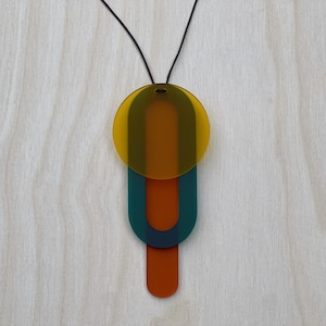 May include: Abstract pendant necklace with a black cord. The pendant is made of translucent acrylic and features a yellow circle, a teal U-shape, and an orange rectangle. The necklace is a modern geometric design.