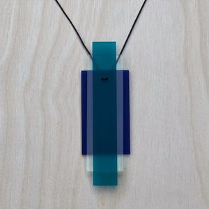 Op de afbeelding: Een moderne hangerketting met een geometrisch ontwerp. De hanger bestaat uit gelaagde rechthoekige vormen in tinten van teal, blauw en helder acryl, hangend aan een zwart koord. De ketting wordt getoond tegen een lichte houtnerf achtergrond.
