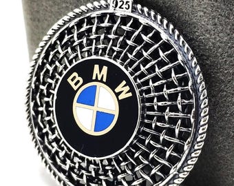 May include: Silver pendant with a woven pattern and a BMW logo at the centre. The logo displays the BMW letters in gold and white, with a blue and white emblem. The pendant is circular and has a textured border.