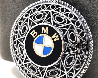May include: A silver pendant featuring the BMW logo. The pendant has an elaborate filigree pattern surrounding the logo, with the number "925" stamped at the top. The BMW logo has a black background with the BMW emblem in blue, white, and gold.