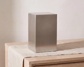 May include: A rectangular, brushed silver-toned metal box with a smooth, flat top and vertical sides. The object features a clean, unadorned surface with a metallic finish, standing upright as a solid, singular, and geometric container.