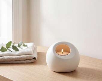 May include: A smooth, matte white ceramic candle holder with a rounded, pebble-like shape and a circular opening. Inside the hollow center sits a small, lit tea light candle with a visible flame, casting a warm glow from within the structure.