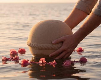 May include: A spherical beige urn with a textured surface and a decorative rope-like band around the center. The urn is partially submerged in water, surrounded by scattered pink flower petals, reflecting the warm light of a sunset.