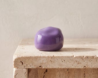 May include: A smooth, rounded, solid purple object with a glossy finish. The item has an irregular, organic shape that resembles a polished stone or a small sculpture. It features a reflective surface that catches light, creating subtle highlights.