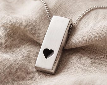 May include: A silver-toned rectangular pendant necklace featuring a small, dark heart-shaped cutout near the bottom. The pendant has a smooth, brushed metallic finish and hangs from a matching silver-toned metal chain link necklace.