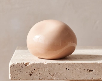 May include: A smooth, rounded, peach-colored ceramic object with a glossy finish. The item has an irregular, organic shape that resembles a polished stone or a soft, rounded form, featuring a reflective surface that catches the light.