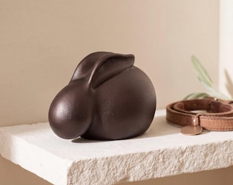 May include: A smooth, dark brown ceramic figurine shaped like a resting rabbit with rounded features and long ears folded back. The sculpture has a matte finish and is positioned next to a tan leather collar with a small circular metal tag.