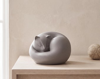May include: A smooth, matte gray ceramic sculpture of a cat curled into a tight, circular sleeping position. The figurine features simple, clean lines with subtle indentations for the ears and closed eyes, creating a solid, rounded form.